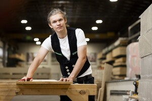 Das Bild zeigt einen jungen Zimmerer, der mit Bleistift und Winkel ein Maß anreißt. Er stützt sich auf sein Werkstück und lächelt in die Kamera.