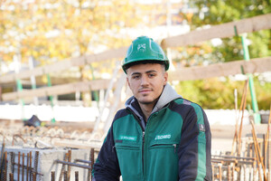 Das Bild zeigt einen jungen Bau-Facharbieter in SCHMID Arbeitskleidung. Im Hintergrund ist eine Baustelle zu erkennen. Er schaut mit einem Lächeln in die Kamera.