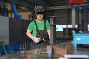 Das bild zeigt einen Metallbauexperten mit einem Schweißbrenner in der Hand. Er steht hinter einem Schweißtisch und trägt einen Schutzhelm mit hochgeklapptem Schild und lächelt in die Kamera.