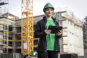Das Foto zeigt eine junge Frau in Arbeitskleidung vor einer Baustelle. Sie hält ein Tablett in der Hand und schaut selbstbewusst in die Kamera.