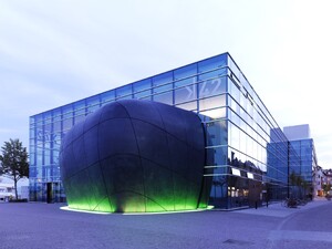 Das bild zeigt einen besonderen Bau. Im Hintergrund einen Kubus aus Glas, im Vordergrund eine rundes Gebilde, das aus dem Kubus ragt. Es handelt sich um das K42 in Friedrichshafen.