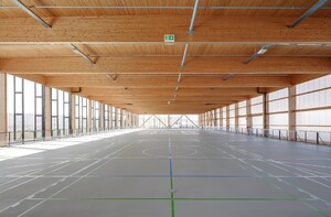 Das Foto zeigt eine große Sporthalle mit Holzdecke und großen Fensterflächen rechts und links der Halle.