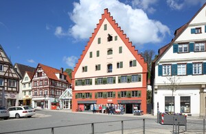Das Foto zeigt eine mittelalterliche Häuserfront. Im Mittelpunkt fällt ein Gibel in oranger und beiger Farbe auf. Im EG befindet sich ein Ladengeschäft. In den oberen Stockwerken scheinen Wohnungen untergebracht zu sein.