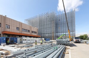 Das Foto zeigt eine Baustelle. Eine riesige Stahlkonstruktion wird montiert. Das spätere Hochregal, das an eine bereits bestehende Logistikhalle anschließt.