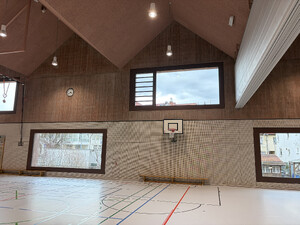 Das Bild zeigt die Turnhalle von innen mit Basketballkorb, Trennwand, großen Fenstern und den roten Wänden