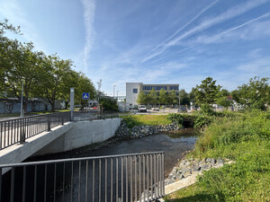 Auf dem Bild sieht man eine Betonbrücke mit Geländer über einen kleinen Fluß, im hintergrund ein Bürogebäude