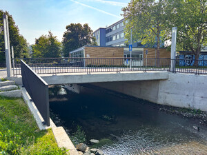 Auf dem Bild sieht man eine neue Brücke mit Geländer, im hintergrund ein Schulgebäude
