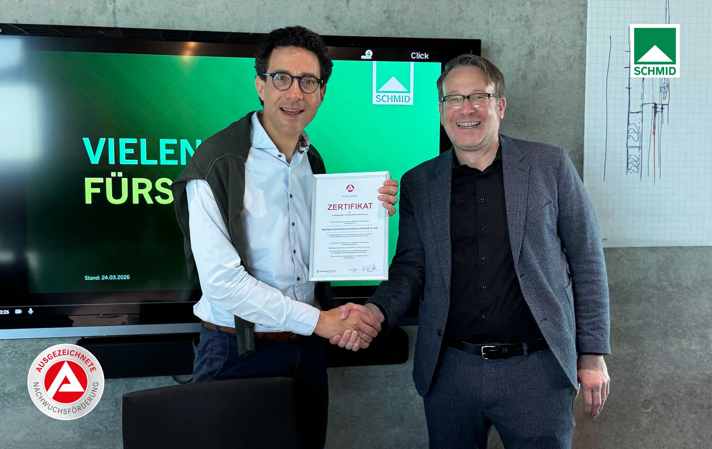 Das Foto zeigt Herrn Dr. Denkmann von der Agentur f&uuml;r Arbeit in Ulm und Herrn Schmid von Matth&auml;us Schmid Bauunternehmen. Herr Denkmann &uuml;berreicht Herrn Schmid das Zertifikat f&uuml;r Ausbildungs- und Nachwuchsf&ouml;rderung. Er gratuliert ihm. Beide l&auml;cheln.