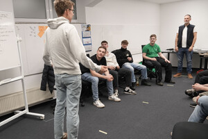 Das Bild zeigt junge Menschen, die im Halbkreis sitzen. Ein Jugendlicher steht vor der Gruppe und sagt etwas. Im Hintergrund steht der Referent Vokram Zschiesche.