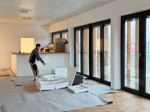 Die Abildung zeigt einen Handwerker, der ein Möbelstück zusammenbaut. Er befindet sich in einem großen Raum mit Bodentiefen Fenstern. Vor den Fenstern laufen die Fassadenteile aus Cortenstahl.