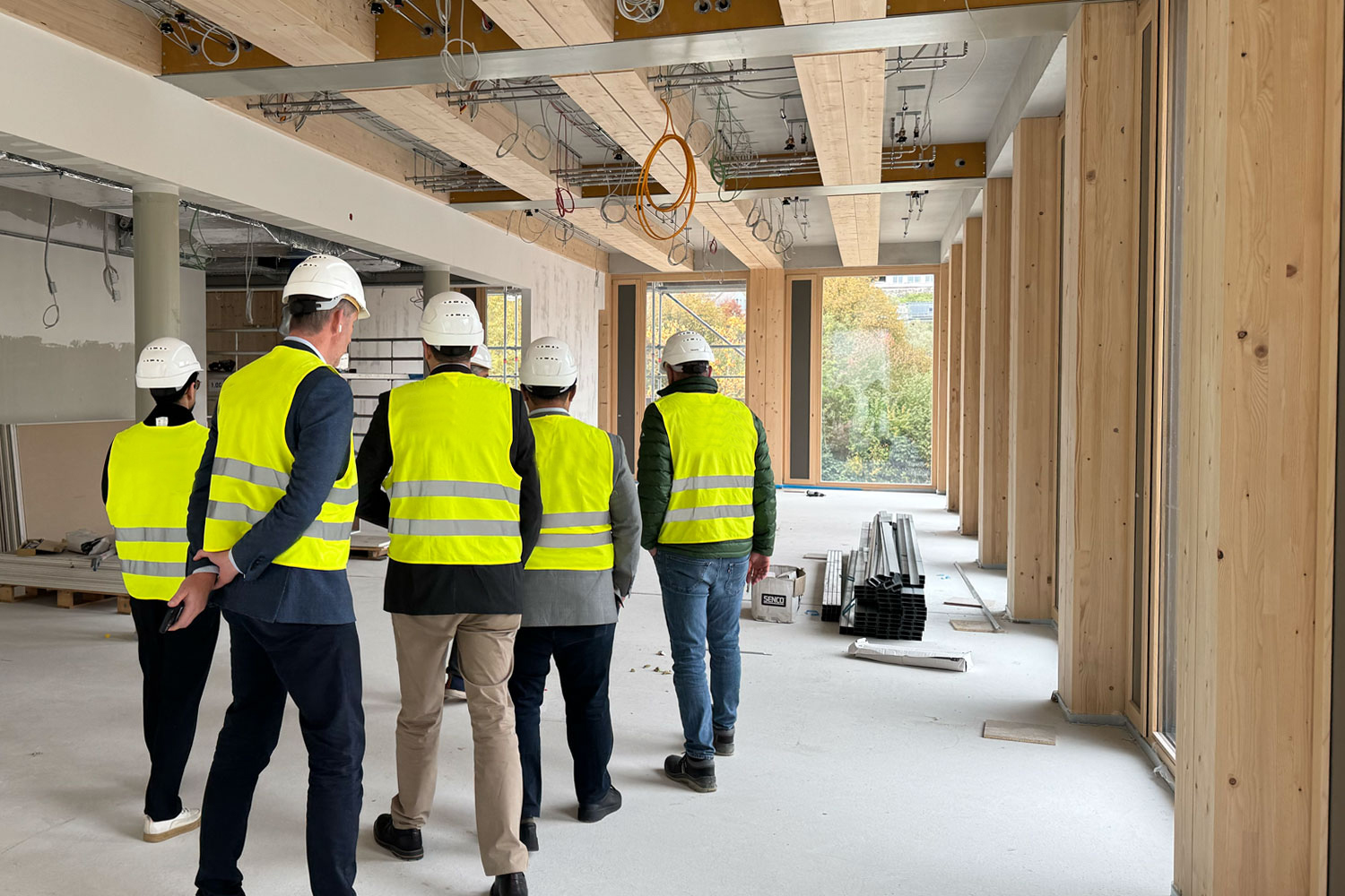 Das Foto zeigt eine Besuchergruppe in Sicherheitswesten und Bauhelmen, die sich die Baustelle von innen anschauen. Man sieht die Holzkonstruktion des Innenraumes.