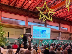 Das Foto zeigt die Geschäftsführer Felix, Christian und Fridolin Schmid auf der Bühne. Im Hintergrund läuft eine Powerpointpräsentation ab. Gezeigt werden Bauprojekte aus dem Jahr 2025. Im Vordergrund sitzen Schmid Mitarbeitende.