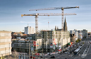 Auf dem Foto ist eine große Baustelle und 2 Krane zu sehen. Im Hintergrund ist das Ulmer Münster zu sehen. Die Baustelle ist im Rohbauzustand und das bestehende Gebäude eingerüstet