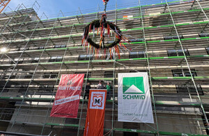 Auf dem Bild ist ein großes eingerüstetes Gebäude zu sehen. Vor dem Gebäude wird ein Richtkranz an einer Kette mittels Kran hochgezogen. Auf dem Gerüst stehen Handwerker, die den Richtspruch sprechen