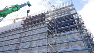 Das Foto zeigt einen großen eingerüsteten Rohbau. Gerade wird mit einem hohen Autokran Baumaterial hochbefordert.