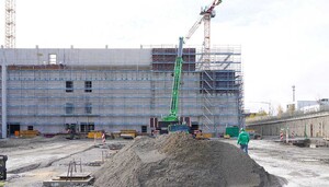 Das Bild zeigt eine Baustellenansicht. Im Vordergrund einen Sandhaufen, im Hintergrund ein großes Stahl-Betongebäude in der Rohbauphase.