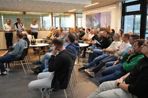 Das Foto zeigt sitzendes Publikum im Atrium des SCHMID Verwaltungsgebäudes. Das Publikum hört einen Vortrag der Geschäftsleiter, die seitlich an der Wand stehen. Es wird einen PPT-Präsentation abgespielt.