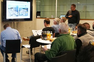 Das Foto zeigt sitzendes Publikum im Atrium des SCHMID Verwaltungsgebäudes. Das Publikum hört den Vortrag eines Bauleiters, der seitlich im Hintergrund steht und spricht. Es wird einen PPT-Präsentation abgespielt.