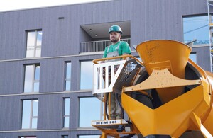 Das Bild zeigt einen Baufacharbeiter, der auf der Brücke des Betonmischers steht und diesen steuert.