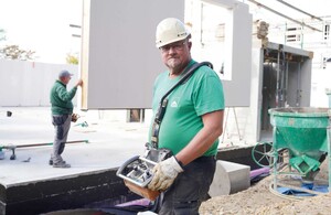 Das Foto zeigt im Vordergrund einen Kranführer, der in die Kamera schaut. Im Hintergrund wird ein Fertigbauelement abgesetzt. Ein weiterer Facharbeiter koordiniert das Absetzen des Bauteiles.