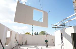 Das Foto zeigt, wie ein Fertigbauelement (Holzständerbauweise) per Kran über der Baustelle eingehoben wird. Auf der Bodenplatte steht der Kranführer, der den Einhob steuert.